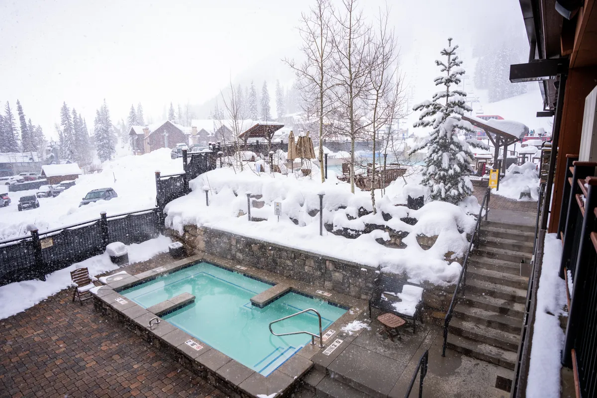 A serene outdoor rooftop hot tub surrounded by a snow-covered landscape, with trees in the background and a staircase leading up. The scene captures inviting, steamy water contrasting with the winter chill, evoking a sense of exclusive relaxation and luxury.