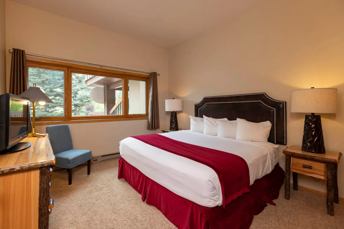 A serene bedroom featuring a king-size bed with crisp white linens and bold red accents. Natural light streams in from a large window, illuminating the decorative lamps and cozy reading nook. The tranquil decor and plush bedding evoke a sanctuary ideal for rest after outdoor adventures.