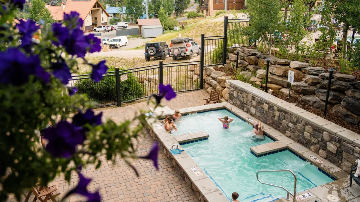 A vibrant outdoor swimming pool bordered by blooming flowers, with children playing and a stone wall enclosing the area. The setting captures joyful poolside moments and the pleasures of a well-maintained resort pool.