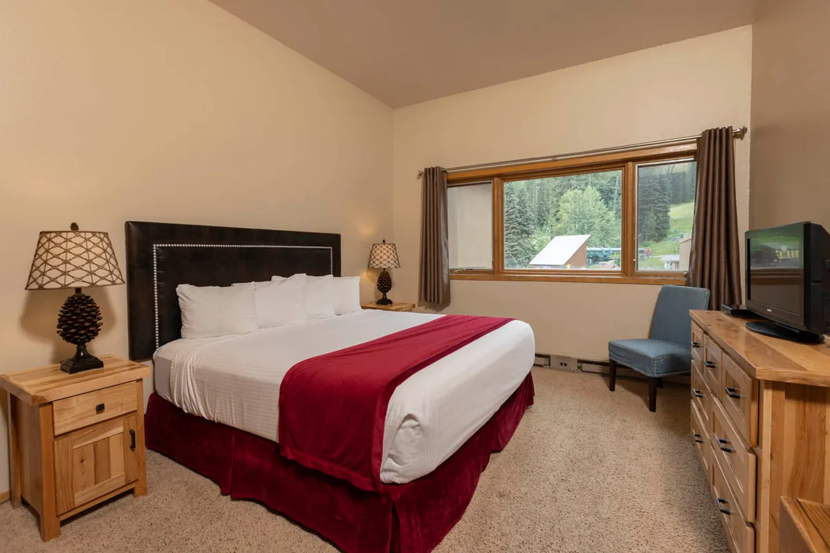 Another elegant bedroom with a king-sized bed, dressed in white linens accented by red details. Wooden nightstands with lamps flank the bed, while a large window offers unobstructed mountain views, enhancing the room’s peaceful ambiance.