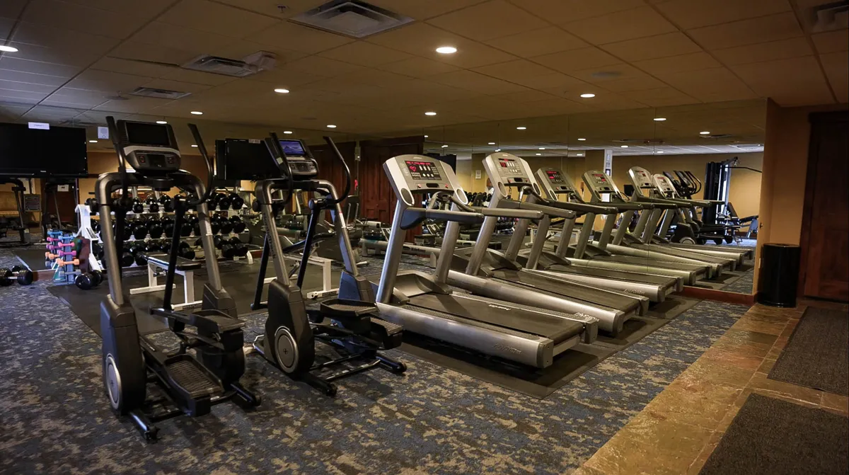 A bright, modern fitness center interior featuring elliptical machines, treadmills, dumbbells, and large mirrors. The setting is motivational and well-equipped, ready for guest workouts and group fitness sessions.