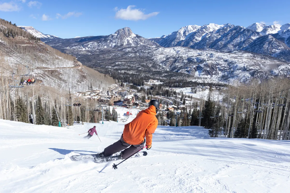 A dynamic view featuring skiers in colorful jackets racing down pristine, snow-covered mountain slopes with the ski resort and breathtaking peaks in the background under a clear blue sky. The sense of motion and camaraderie among the skiers captures the excitement of alpine sports.