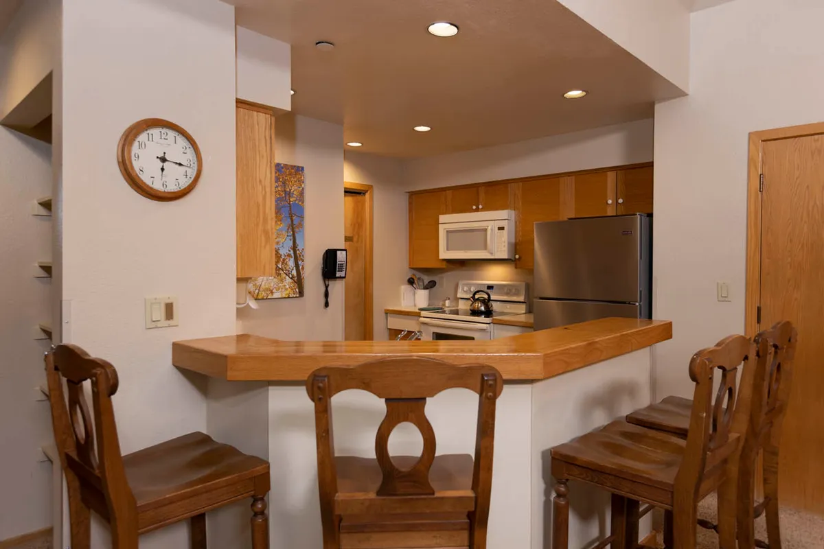 A cozy kitchen scene featuring a breakfast bar, modern kitchen appliances, and warm wooden cabinetry. The space is bathed in morning light, and the presence of a wall clock and inviting countertop suggest a welcoming atmosphere for cooking or casual dining. The layout encourages both meal preparation and social interaction.