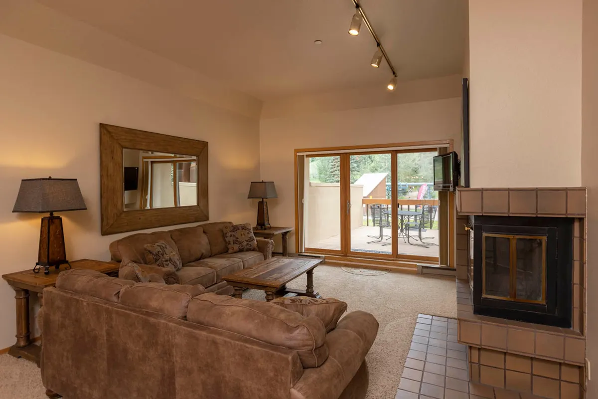 A welcoming living space with a comfortable sofa, elegant coffee table, and a fireplace set against a backdrop of a large mirror and sliding glass doors. The doors open onto a patio, extending the living area into the mountain landscape and bathing the room in natural light.