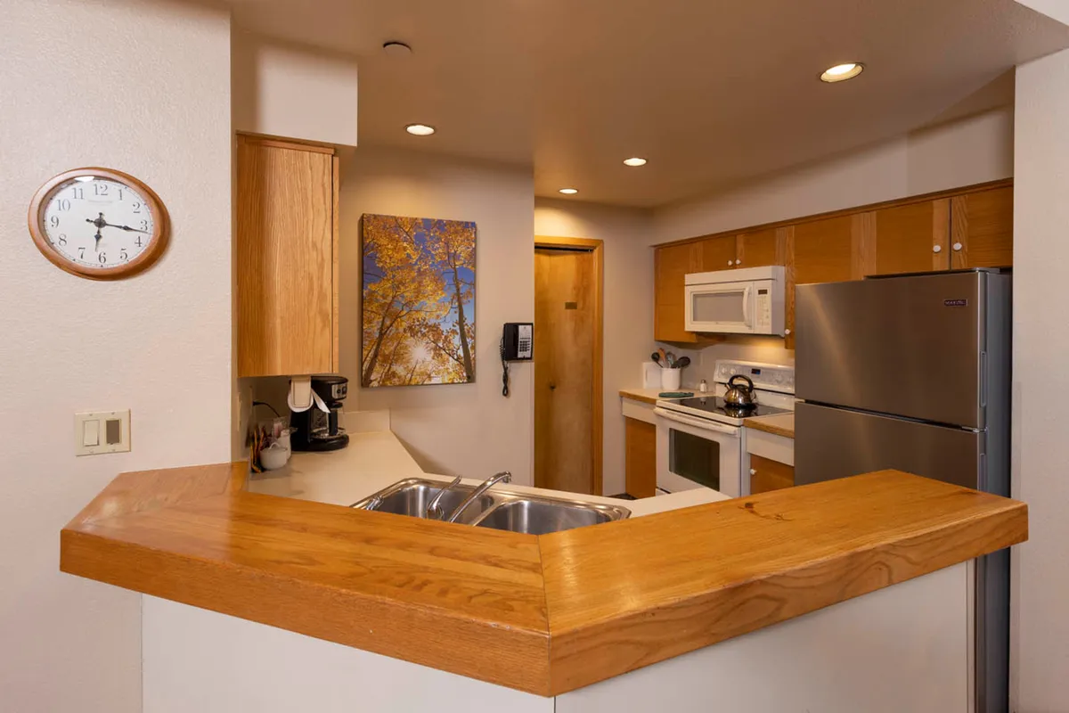 A view of the kitchen and adjacent dining area, centered around a wooden counter and complemented by contemporary appliances and wall art. The setup is designed for group meals and festive gatherings, with a clear emphasis on functionality and sociability. Natural light enhances the cozy, inviting aura of the space.