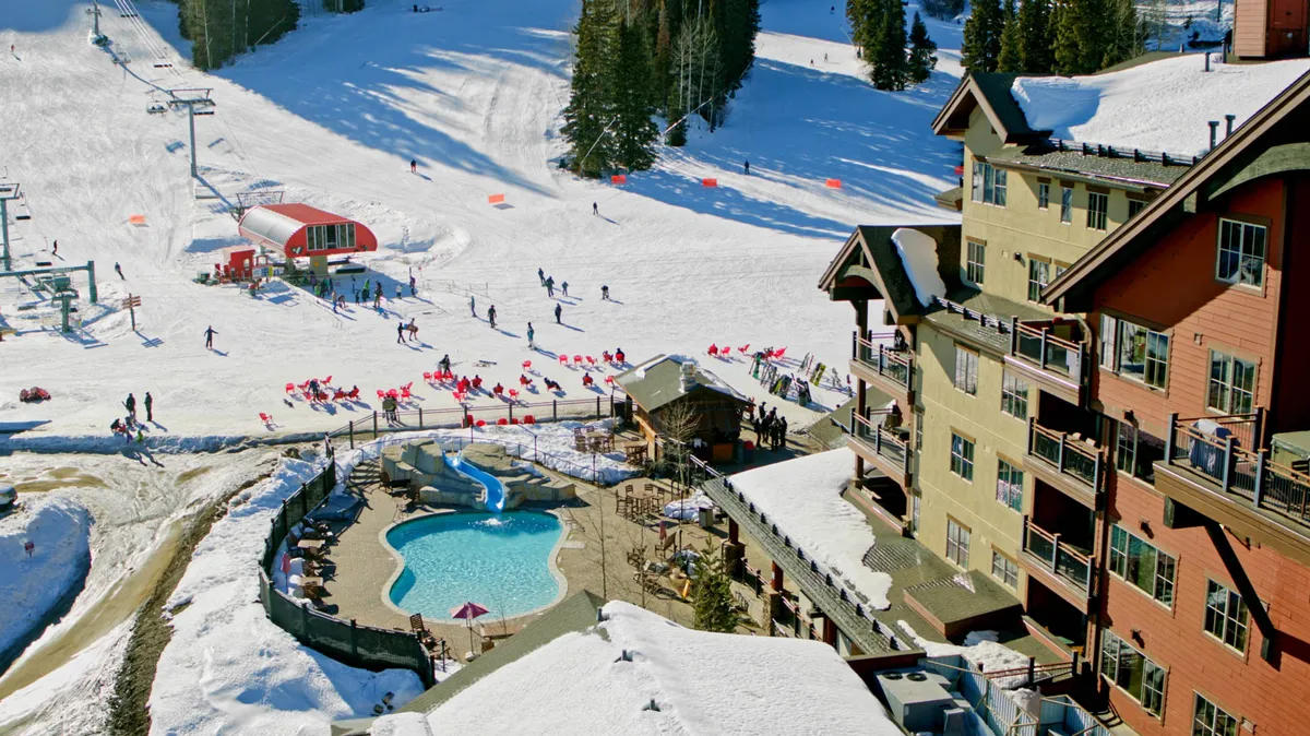 An energetic winter scene showing active ski slopes, a sparkling swimming pool, a lodge, and families enjoying both the snow and warm water. The juxtaposition of skiing and swimming highlights a range of activities available at the resort, from thrilling runs to relaxing dips and cozy time at the lodge.