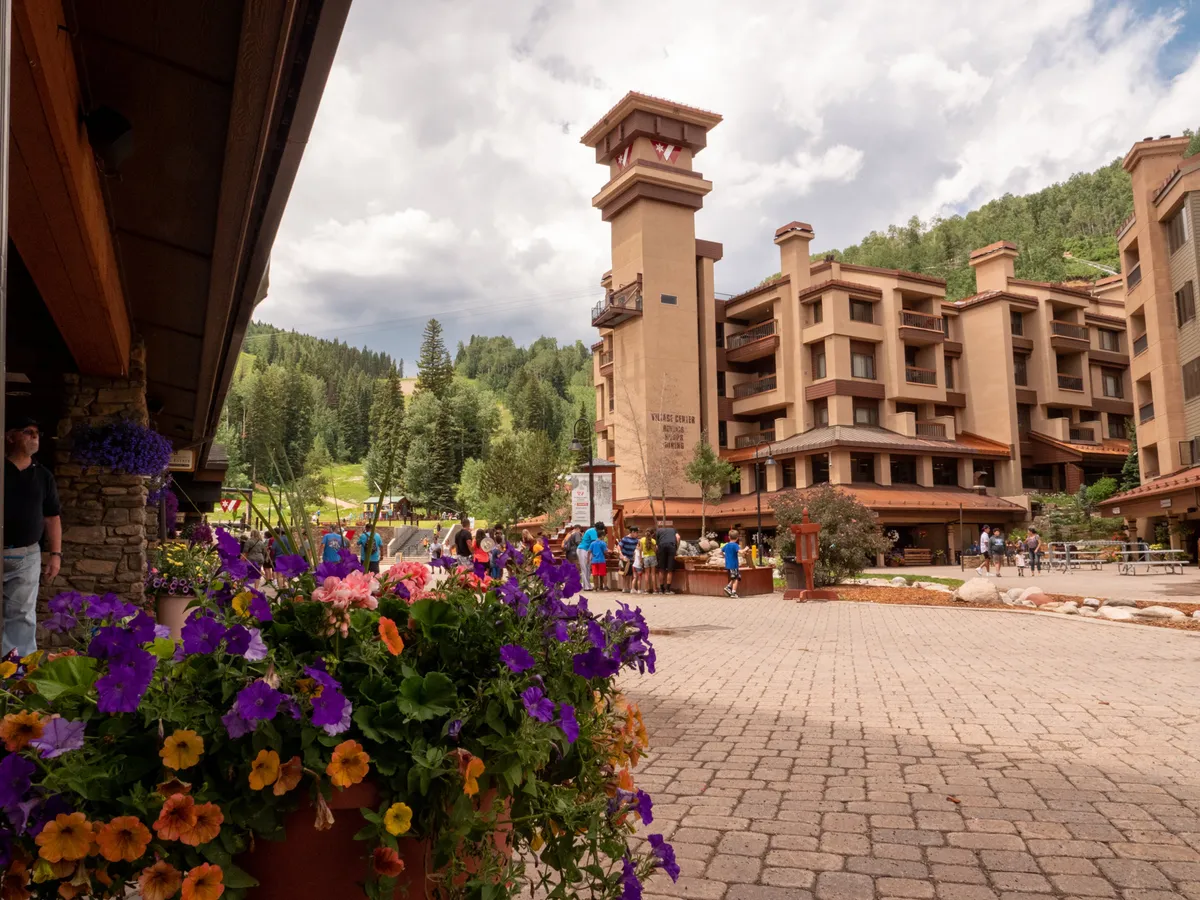 This image showcases the exterior of the Village Center building with its distinctive clock tower, vibrant flower pots, and people enjoying the lively outdoor space. The scene hints at a welcoming community atmosphere, with areas for socializing and relaxing against the backdrop of the resort.