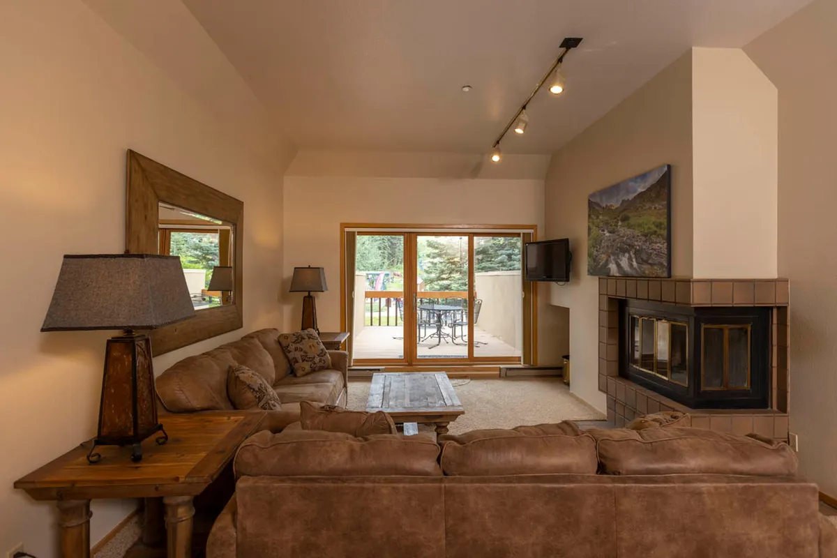 This image showcases a cozy living room with a plush sofa oriented toward a stone fireplace. Large sliding glass doors lead out to an outdoor patio, allowing natural light to flood the room and offering clear sightlines to the mountainous landscape beyond. The space is designed for relaxation and socializing, with warm textures and inviting decor.