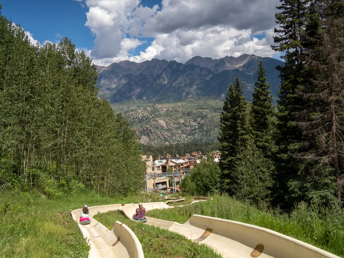 A joyful summertime scene with families and children sliding down a hillside alpine slide surrounded by green mountains, trees, and blue skies. People are clearly engaged in outdoor, family-friendly activities, capturing the playful spirit of the warmer seasons.